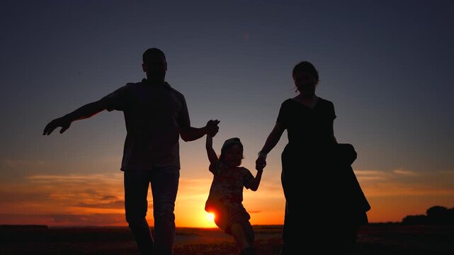 A man and a woman hold hands, silhouetted against a sunset. A child is joyfully lifted by the woman, creating a playful scene near the horizon.