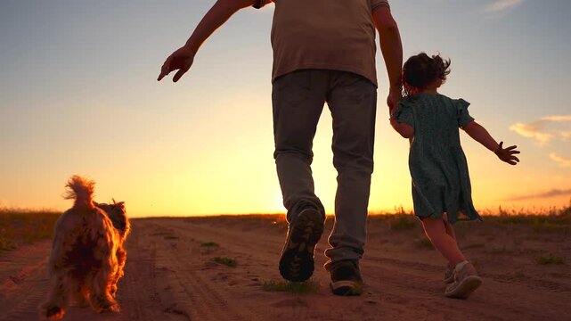 A man and a girl walk hand in hand along a dirt path at sunset. A dog follows closely as they enjoy the warm evening light, creating a serene atmosphere.