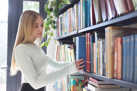 Smart teenage girl picking literature from shelves, enjoying reading and studying in a library or bookstore. 