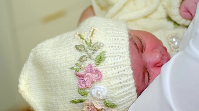 The camera lingers on a cream knitted baby bonnet with floral details as a newborn rests quietly in maternal arms. This scene fits tenderness, protection, handmade clothing, and early bonding.