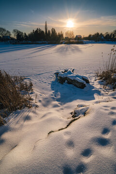 tunning winter landscape at sunset with a frozen, snow-covered lake. Long dramatic tree shadows stretching across the ice, with a snow-dusted log and footprints in the foreground under a bright sunbur