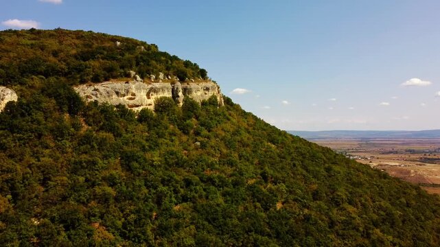 Aerial tracking shot of forested cliffside gliding past open valley revealing layered limestone outcrops under clear afternoon sky