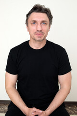 Obraz premium middle aged man with blue eyes and stubble in black t-shirt sitting with hands clasped looking at camera, white wall background portrait, headshot