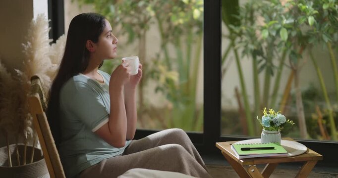Indian calm young adult gen z relax woman sitting on chair pick up cup drink coffee enjoy day room girl hold pen book writing new work notes plan idea spend time take casual job break home house place