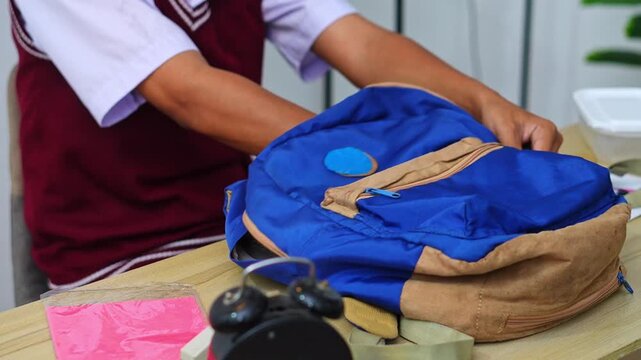 Student in maroon uniform packing stack of books and leather binder into backpack on wooden desk
