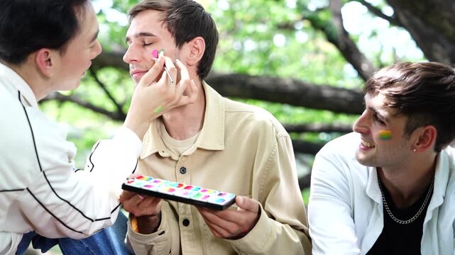 Pride and LGBTQ concept. LGBTQ men sit having a picnic, painting rainbow flags on their friends' faces and smiling happily together in the park.