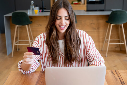 Woman using credit card and laptop at home kitchen table making an online payment and shopping