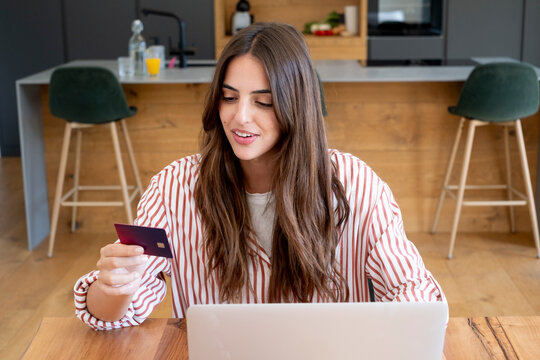 Young woman paying bills and shopping online with a credit card at her laptop in a kitchen