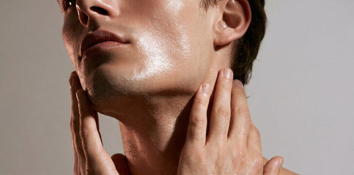 Primer plano del rostro y cuello de un hombre con piel h&uacute;meda y brillante toc&aacute;ndose con las manos. Concepto de rutina de cuidado de la piel masculina, hidrataci&oacute;n y cosm&eacute;tica natural