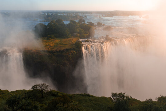 Aerial sunset view of Victoria Falls, Zimbabwe, shows Zambezi River plunging over basalt cliff, mist rising, islands and channels at the brink, and gorges veiled by spray.