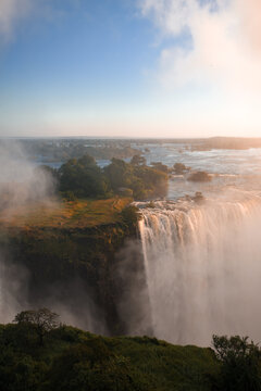 Aerial view of Victoria Falls at sunset on the Zimbabwe side, Zambezi River surges over basalt cliff into misty gorge, tree lined islands and golden spray illuminate deep chasm.