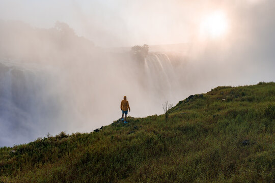 A person in a yellow jacket stands on a grassy cliff in Zimbabwe, gazing at misty Victoria Falls as warm light breaks through, spray rises from the Zambezi River gorge.