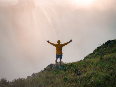 A traveler in a yellow rain jacket stands on a grassy cliff near Victoria Falls, Zimbabwe, arms outstretched toward golden mist as warm light and soft haze suggest roaring water.
