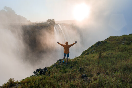 An adventurer in an orange jacket stands on a grassy cliff, arms outstretched toward Victoria Falls horseshoe edge, thunderous spray and warm sunset haze on the Zimbabwe side.
