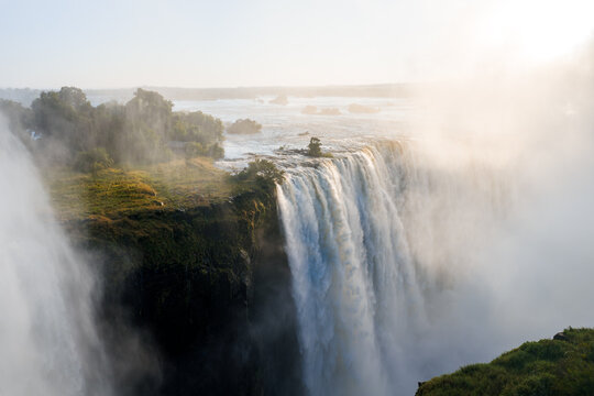 Mist rises over Victoria Falls as the Zambezi plunges over basalt into a shadowed gorge in Zimbabwe, warm low light gilds islands and edges, haze softens horizon.