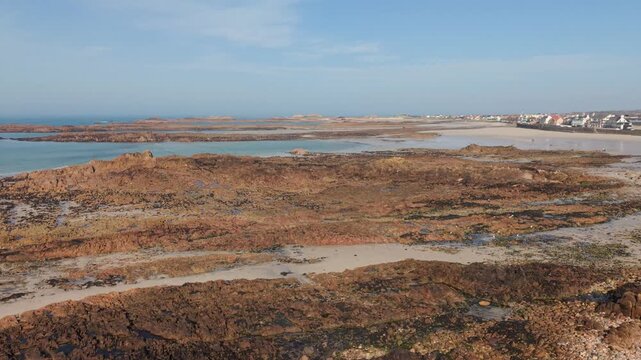 Cobo Bay Guernsey Channel Islands forward drone flight over pink granite rock formations with golden sandy beaches calm sea and coastline in late afternoon sun with blue sky and coastal views