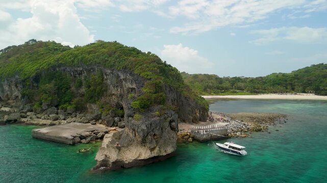 Aerial orbit around Cujo's Keyhole limestone headland at Crystal Cove, Boracay, with tourist walkway along cliff base, white catamaran boat, and turquoise waters, Philippines