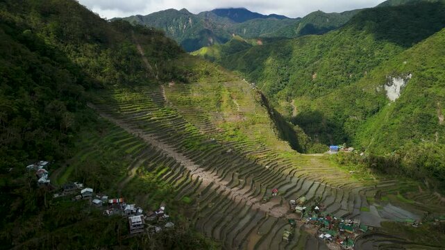 High-angle aerial parallax above Batad Rice Terraces in Banaue reveals stepped terraces in mountain bowl with two Ifugao villages, deep valley river and Cordillera peaks