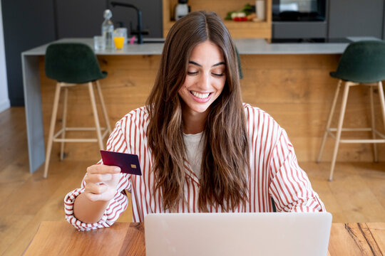 Young woman smiling while using credit card and laptop for online shopping at home kitchen table