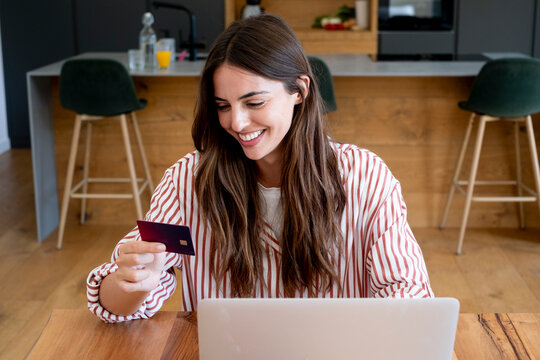 Smiling woman paying with a credit card on a laptop in a modern kitchen at home