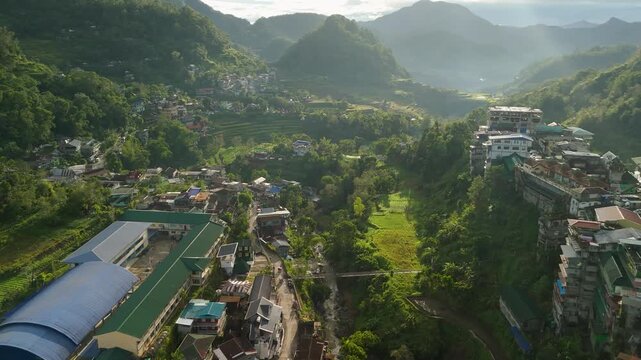 Sweeping aerial flyover reveals Banaue town tucked in lush green mountain valley of Ifugao, Philippines, with rice terraces, misty peaks and clustered buildings