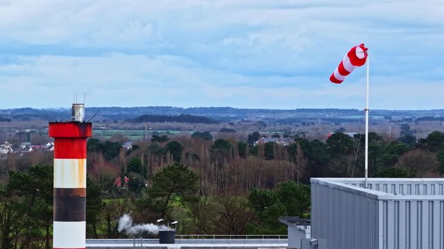 A red and white chimney stands beside a windsock on a rooftop overlooking a rural landscape. Ideal for use in industrial or environmental documentaries.