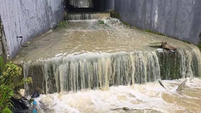 Polluted wastewater discharging from a concrete channel into a river, creating white foam downstream and impacting the environment, symbolizing water pollution and ecological damage