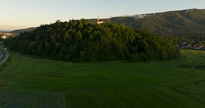 View of St. Marietta church on Pekrska Hill near Maribor, Slovenia. Forested hill, green countryside and Pohorje Mountains illuminated by warm spring evening light.