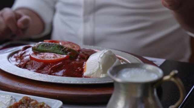A man in a white shirt enjoys a traditional Turkish Iskender kebab at a restaurant. Close-up shot showing meat, yogurt, and grilled vegetables on a metal tray with a shallow depth of field.