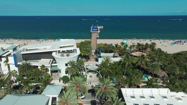 Aerial drone view of the iconic Pompano Beach Pier in Florida, stretching into the deep blue Atlantic, framed by palm trees, sandy shores, and the lively oceanfront plaza.