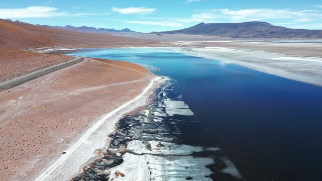 Backward reveal flight over Laguna Tara deep blue waters with jagged salt crust shoreline winding road red desert terrain and Andean peaks under blue sky. Laguna Tara, San Pedro de Atacama, Chile