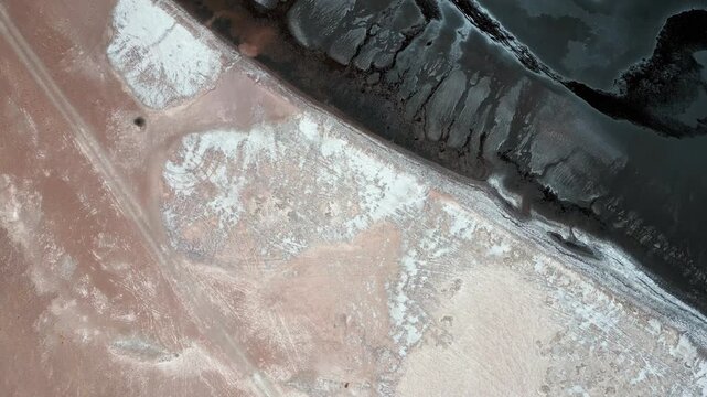 Top down aerial view of Laguna Tara dark water shoreline with abstract white salt crust patterns and sandy sediment creating dramatic natural contrast. Laguna Tara, San Pedro de Atacama, Chile