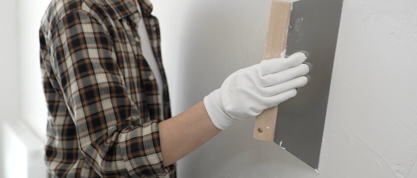 Unknown female construction worker is wearing and checkered shirt is applying plaster with professional grade spatula, smoothing white wall during home renovation project with skilled craftsmanship