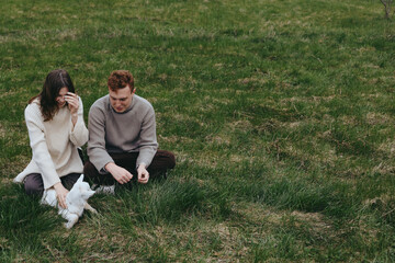 Fototapeta premium Couple sits on grass outdoors sharing a playful moment with a goat kid nearby, creating a warm lifestyle scene focused on companionship and gentle animal interaction in soft light. Cozy.