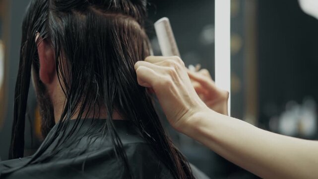 Female barber cutting long wet hair of male client with scissors and comb during professional haircut procedure in modern barbershop