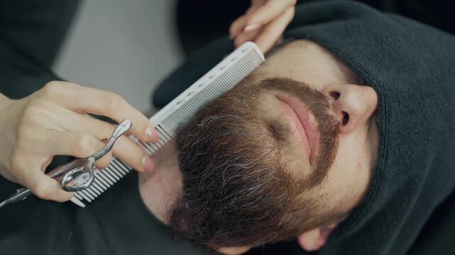 Professional barber trimming beard of male client with scissors and comb during grooming procedure in modern barbershop salon