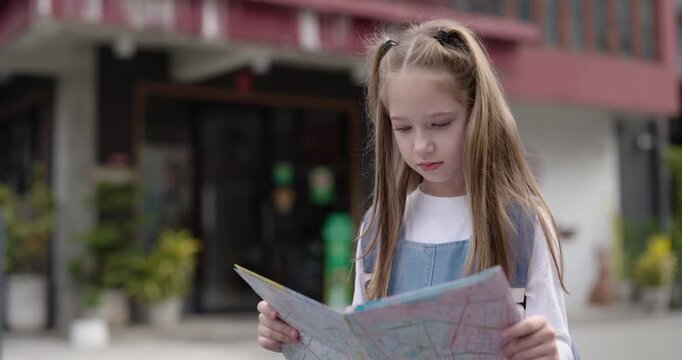 Portrait of focused little girl reads map as stands in historical town district. Dedicated junior schoolgirl studies urban plans to choose route in city center