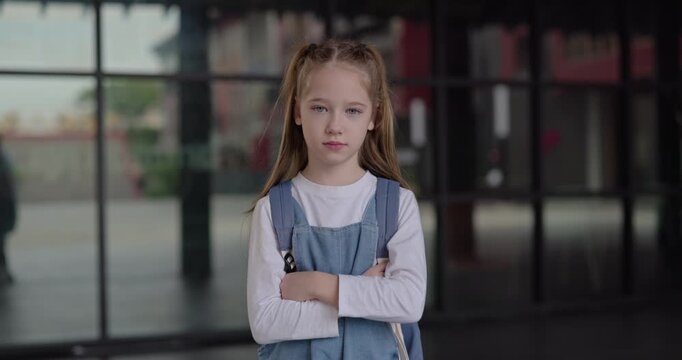 Portrait of calm little girl in denim overall stands near building with glass facade. Junior schoolgirl with crossed arms gives calm expression on city street