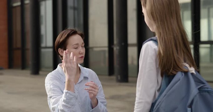 Portrait of mother gestures and talks to little girl student near school building. Caring woman explains behavior rules to little daughter on city street
