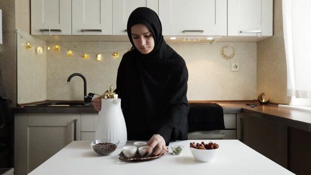 muslim woman in black hijab setting a small finjan cup on tray beside white dallah dates coffee beans and cardamom during home hospitality preparation