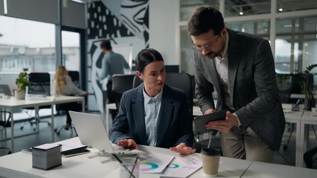 Two Caucasian business colleagues talking discuss data in digital tablet compare information in laptop businesswoman female with businessman male leader talk together analysing online work in office