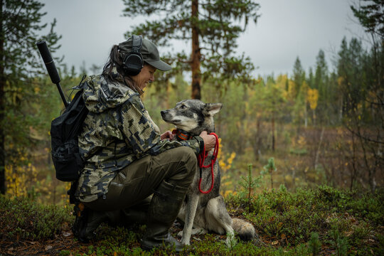 Side view of smiling female hunter petting dog while kneeling on ground in forest