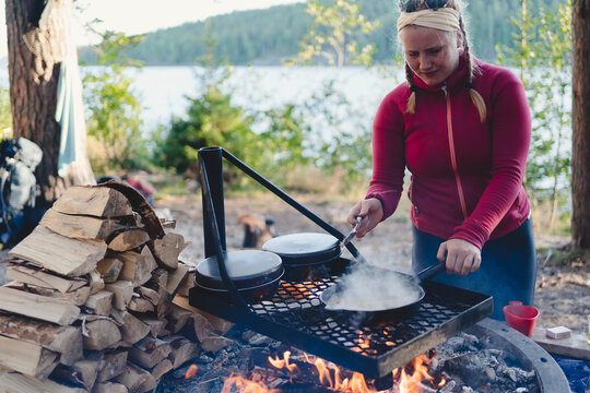 Young female traveler cooking food on fire pit while camping in forest