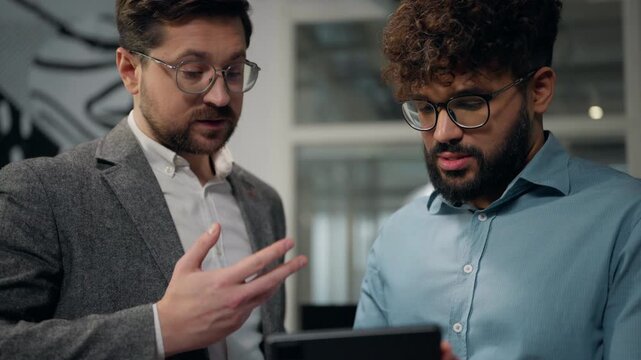Two happy multiracial business men smiling communicating looking in digital tablet cooperate talking teamwork with wireless computer diverse businessman Caucasian man with Arabian guy talk in office