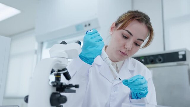 A scientist in a lab uses a pipette to transfer liquid into a petri dish while working at the microscope. The setting is clean and organized with lab equipment around.