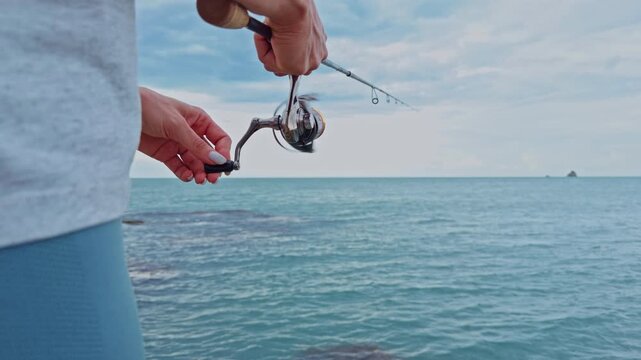 Close View of Young Woman Reeling Spinning Reel Above Calm Tropical Ocean Near Rocky Coastline Under Cloudy Sky, Slow Motion Recreational Fishing