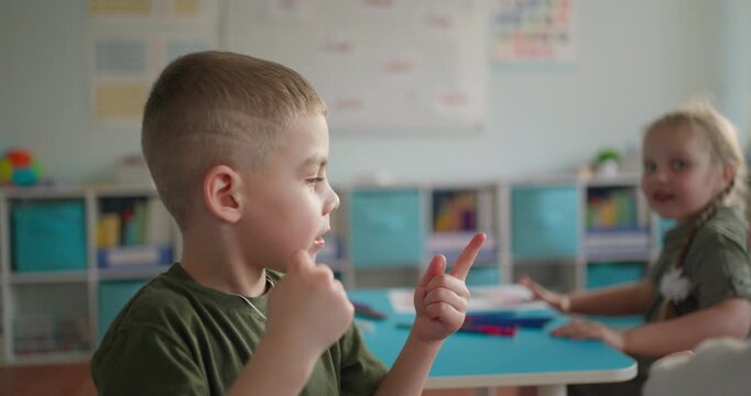 Young boy eating a small snack while sitting with classmates at a classroom table, softly lit educational setting with charts and shelves in background