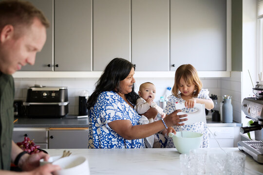 Mother carrying toddler while teaching daughter to use electric beater in kitchen at home