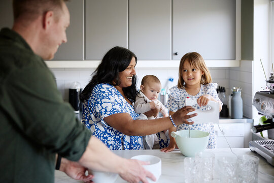 Smiling mother carrying toddler while teaching daughter to use electric beater in kitchen at home