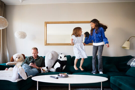 Sisters jumping on sofa while father using smart phone in living room at home